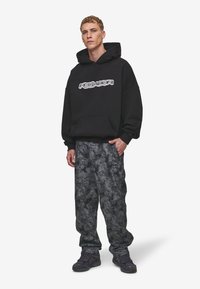 Hanorac negru cu buzunar frontal și logo, asortat cu pantaloni gri de camuflaj, largi. Încălțămintea este întunecată și texturată.