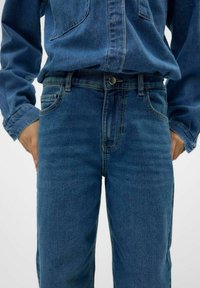 Blå denimjeans med rak passform, försedd med knäppning, framfickor och subtila sömnadsdetaljer. Tyget har en slät textur.