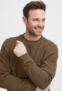 La camicia a maniche lunghe marrone con texture presenta polsini a coste, un colletto a girocollo e una patch con logo sulla manica sinistra.