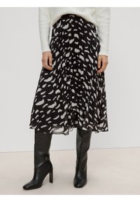 Mujer con falda midi plizada negra con manchas abstractas blancas, suéter de punto blanco y botas altas negras de tacón.