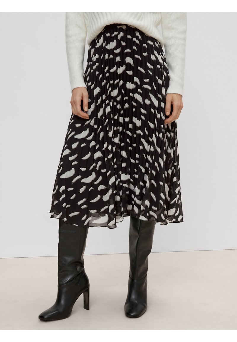 Mujer con falda midi plizada negra con manchas abstractas blancas, suéter de punto blanco y botas altas negras de tacón.
