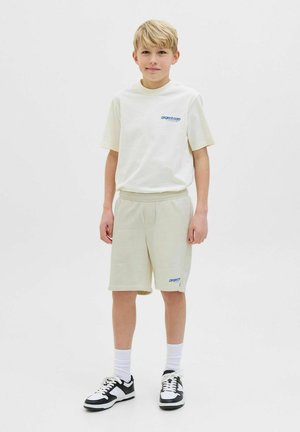T-shirt beige clair avec un logo bleu, associé à un short assorti. Des chaussettes blanches et des chaussures de sport noires avec des accents blancs complètent la tenue.