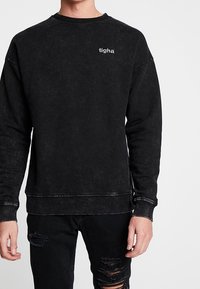 Felpa nera dal taglio comodo, con un piccolo logo bianco sul petto, collo a costine e dettagli di cuciture visibili.