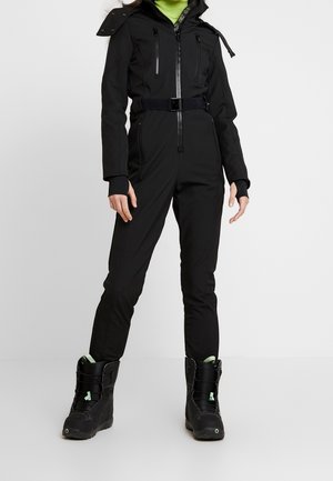 Osoba ubrana w czarny kombinezon zapinany na zamek z paskiem oraz czarne zimowe buty z miętowymi akcentami stoi na jednolitym białym tle.