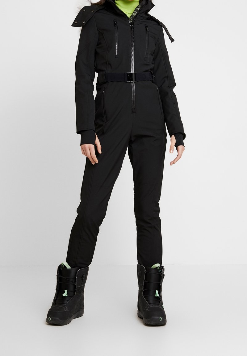 Personne portant une combinaison noire zippée avec une ceinture et des bottes d'hiver noires avec des accents vert menthe, se tenant sur un fond blanc uni.