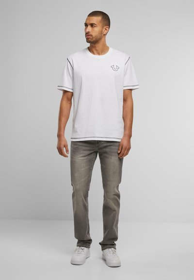 T-shirt blanc à manches courtes avec un logo sur la poitrine, associé à un jean gris clair et des baskets blanches, avec des coutures contrastantes sur les manches.
