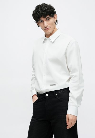 Ung mand med krøllet hår iført en hvid skjorte med knapper og sorte jeans, stående med den ene hånd i lommen mod en ensfarvet baggrund.