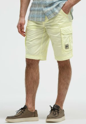 Mann trägt hellgelbe Cargo-Shorts, ein kariertes Hemd und braune Freizeitschuhe, steht mit einer Hand in der Tasche.