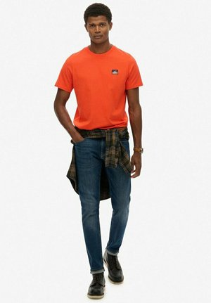 Orange bomulds T-shirt med et lille logo, parret med blå denim jeans og sorte læder støvler. En ternet skjorte bundet om livet.