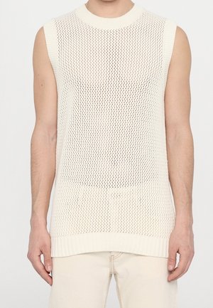 Männlich, trägt einen ärmellosen, cremefarbenen Strick-Netzpullover über hellen Hosen, steht vor einem schlichten, hellen Hintergrund.