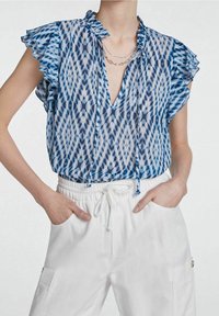 Blouse transparente à motifs bleus et blancs avec manches courtes à volants et un profond col en V, associée à un pantalon blanc à cordon de serrage.