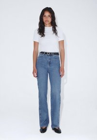 Camisa blanca de manga corta, jeans acampanados de cintura alta azul con bolsillos traseros y cinturón negro. Pose de pie, zapatos negros planos. Fondo neutro.