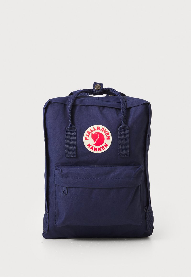 Mochila azul marinho com forma retangular, feita de tecido durável, com um bolso frontal e um patch de logótipo circular. Duas alças superiores.