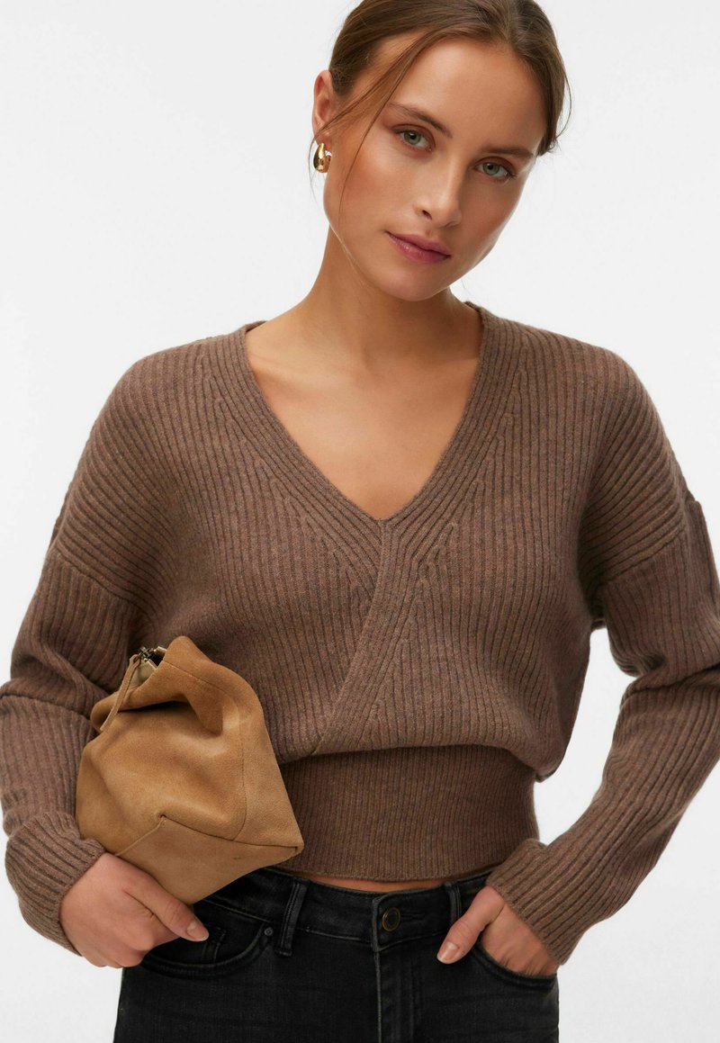 Femme portant un pull marron à côtes avec col en V et un jean noir, tenant un sac à main en daim beige sur fond blanc