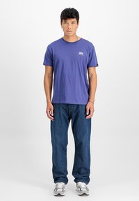 Violetas kokvilnas T-krekls ar īsām piedurknēm un apaļu kakla izgriezumu, kombinēts ar ziliem džinsiem un pelēkām kedām ar baltu zoli.