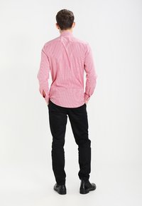 Maglietta a righe rosse e bianche a maniche lunghe con colletto button-down, abbinata a pantaloni neri e scarpe nere, vista da dietro.