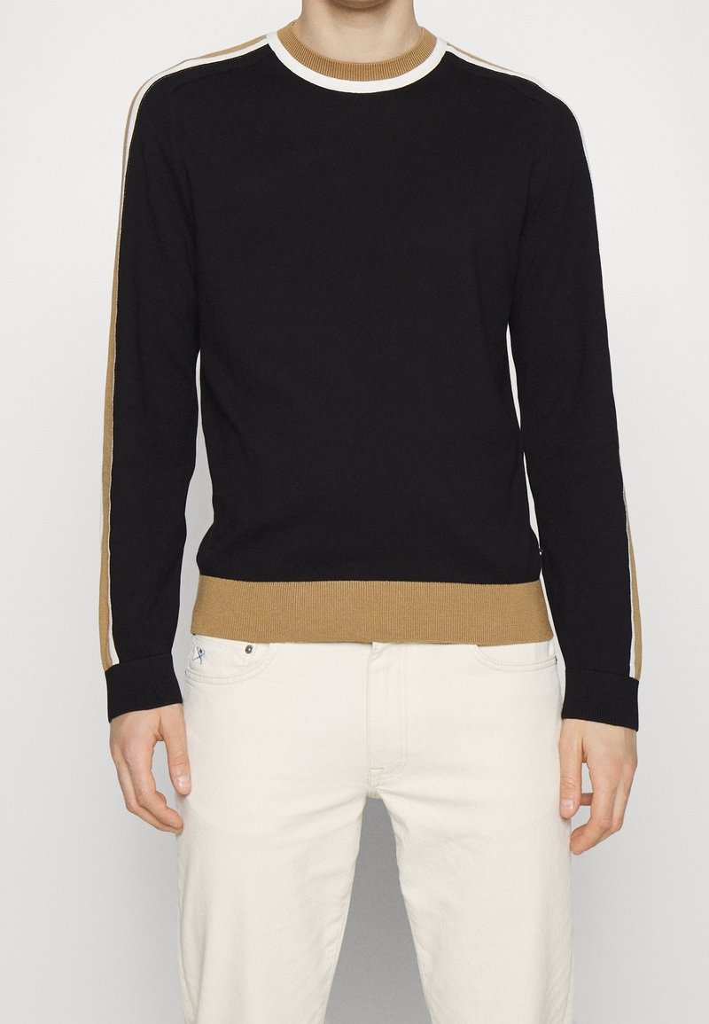 Pull en maille noire avec des rayures camel et blanches sur les manches, col rond et ourlet côtelés, coupe ajustée, porté avec un pantalon clair.