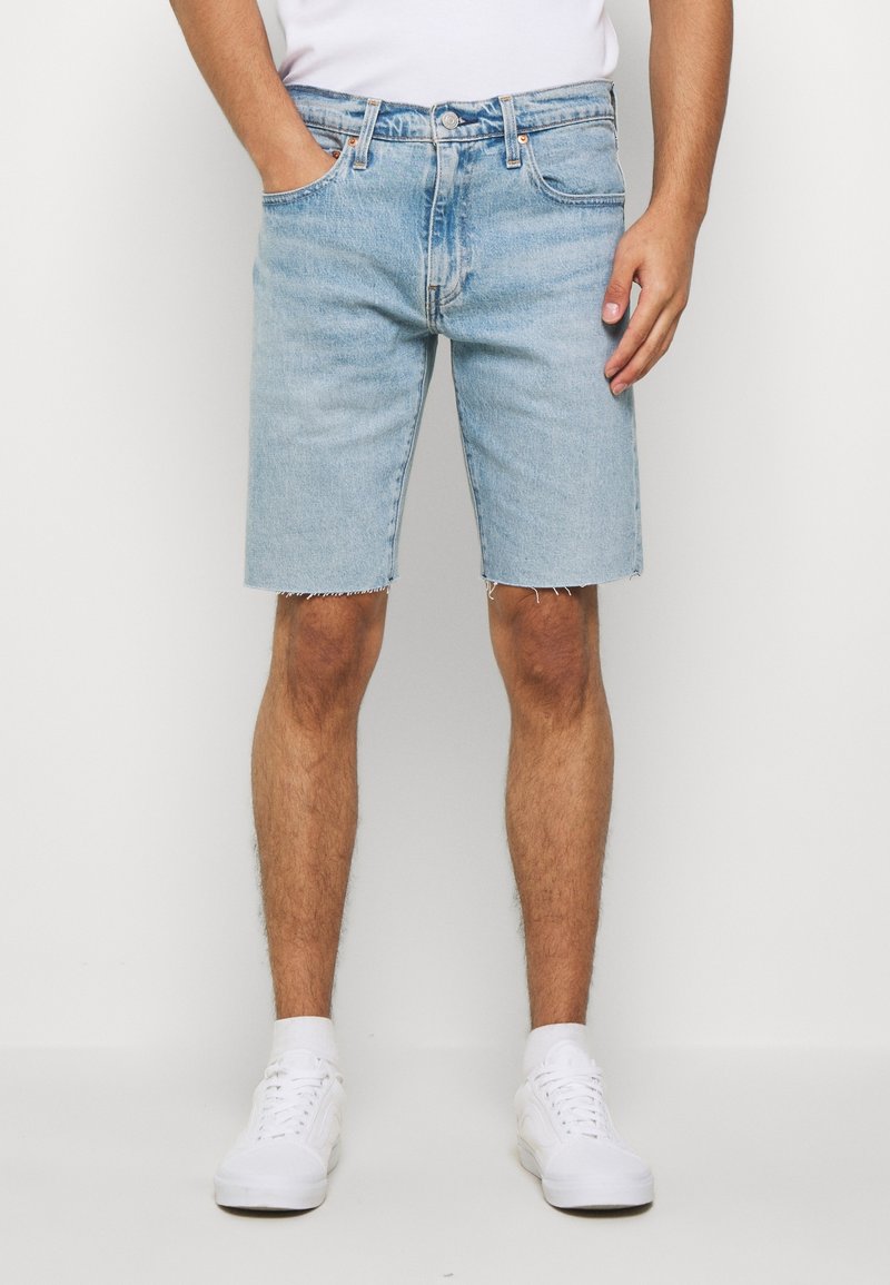 Hombre vistiendo pantalones cortos de mezclilla azul claro con dobladillo sin terminar, calcetines blancos y zapatillas blancas, de pie contra un fondo blanco liso.