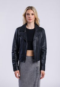 Černá kožená zkrácená bunda s širokým límcem, předním zipem a dlouhými rukávy. Nositá přes černý top a šedou sukni s knoflíky.