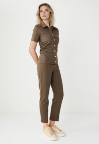 Bruine aansluitende top met korte mouwen en gouden knopen, gecombineerd met bijpassende taps toelopende broek en lichtbeige sneakers.