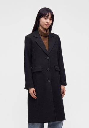 Jeune femme portant un long manteau noir boutonné par-dessus un pull à col roulé marron et un jean bleu, debout devant un fond clair uni.