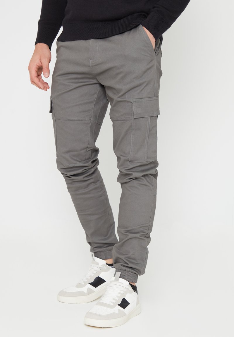 Grijze cargo broek met elastische boorden, meerdere zijzakken en een gladde, licht gestructureerde stof. In combinatie met witte sneakers met zwarte accenten.