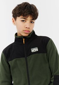 Groene en zwarte fleecejack met hoge kraag, volledige rits en logo-opstrijkpatch. Beschikt over een gladde textuur en contrasterende kleurvlakken.