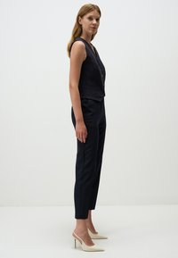 Donker mouwloos vest met een ribbelstructuur en knoopdetail, gecombineerd met tapered zwarte pantalon en crèmekleurige slingback hakken. Gladde stofafwerking.