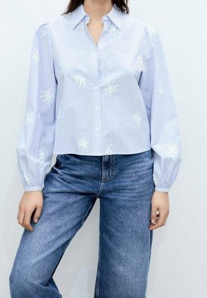 Femme portant un chemisier rayé bleu clair avec une broderie florale blanche et de longues manches bouffantes, assorti à un jean bleu taille haute.