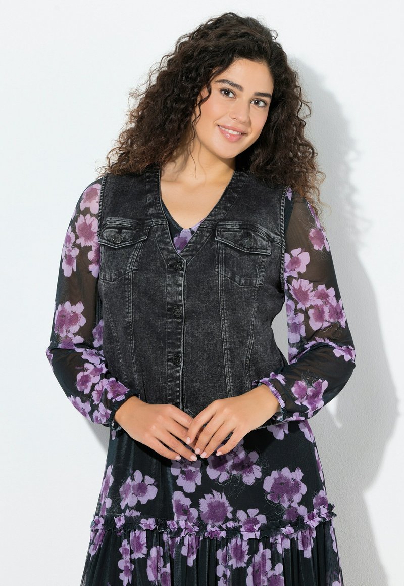Kamizelka dżinsowa z ciemnym wybarwieniem, z przednimi kieszeniami i zapięciem na guziki, noszona na przezroczystej, kwiatowej bluzce w kolorze fioletowym i czarnym.