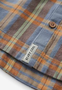 Blaues, oranges, grünes und braunes kariertes Stoff mit einem braunen Knopf und einem weißen Etikett mit der Aufschrift "BEARY COOL", nahe dem Saum eingenäht.