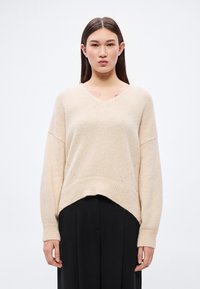 Béžový pletený sveter s výstrihom do V, spustenými ramenami a voľným strihom. Má rebrovaný lem a jemný textúrovaný detail pozdĺž okrajov.