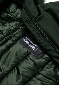 Donkergroene pufferjack met een gequiltde textuur, voorzien van een ritssluiting en een merketiket aan de binnenkant. Het materiaal lijkt glanzend en glad.