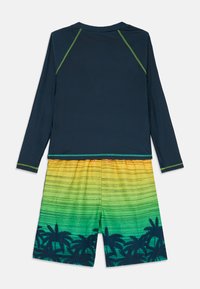Dlhý rukáv rash guard v námorníckej modrej farbe s zelenými akcentmi, v kombinácii s plážovými šortkami s prechodom farieb od žltej po zelenú a siluetou paliem.
