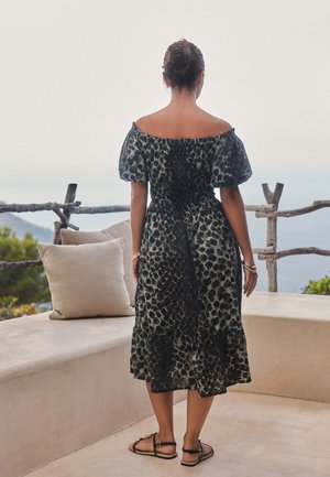 Žena v čiernych a sivých leopardích šatách s odhalenými ramenami a sandále stojí na terase s béžovými vankúšmi a dreveným plotom s výhľadom na prírodu.