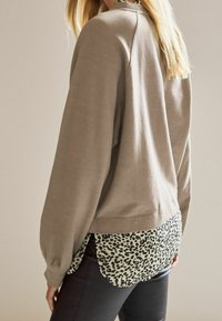 Felpa a maniche lunghe color taupe con orlo curvo, indossata sopra una camicia a stampa leopardata verde e nera. Il tessuto appare morbido e strutturato.