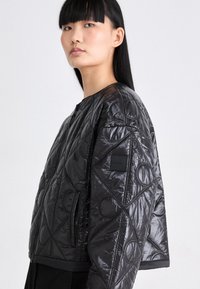 Vrouw met lang zwart haar, gekleed in een glanzende zwarte gewatteerde jas met cirkelvormige stiksel, staande tegen een eentonige lichte achtergrond.