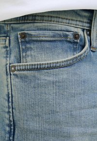 Jean en denim bleu clair avec une poche avant ornée de rivets en métal foncé et de coutures jaunes contrastantes le long des coutures.