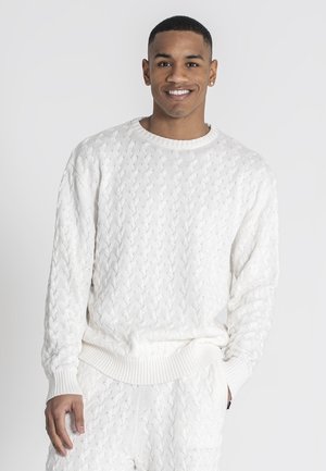 Suéter blanco de punto con un patrón de trenzas texturizadas, que presenta un escote redondo y puños acanalados. El modelo lleva pantalones blancos a juego.