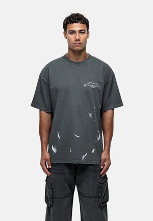 T-shirt in cotone grigio oversize con macchie di vernice bianca vissuta, collo a girocolo a coste, maniche corte e testo del logo in bianco sopra il petto.