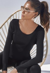 Zwarte lange mouw top met een ronde halslijn, gladde textuur. Model draagt oversized ronde zonnebril en ronde "Love" oorbellen, zittend in een rieten stoel.