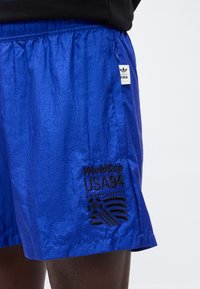 Φωτεινά μπλε αθλητικά σορτς με μαύρο λογότυπο "World Cup USA 94" και ποδοσφαιρική μπάλα κεντημένα στο αριστερό πόδι, φορεμένα με μαύρο τοπ.