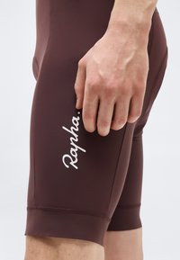 Κοντινό πλάνο ενός χεριού που ακουμπά στον μηρό, φορώντας σκουρόχρωμο καφέ σορτς ποδηλασίας με το λογότυπο "Rapha." στο πλάι.