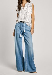 Donna che indossa una blusa bianca senza maniche con ruches e frange, abbinata a pantaloni a gamba larga blu a vita alta e una cintura nera.