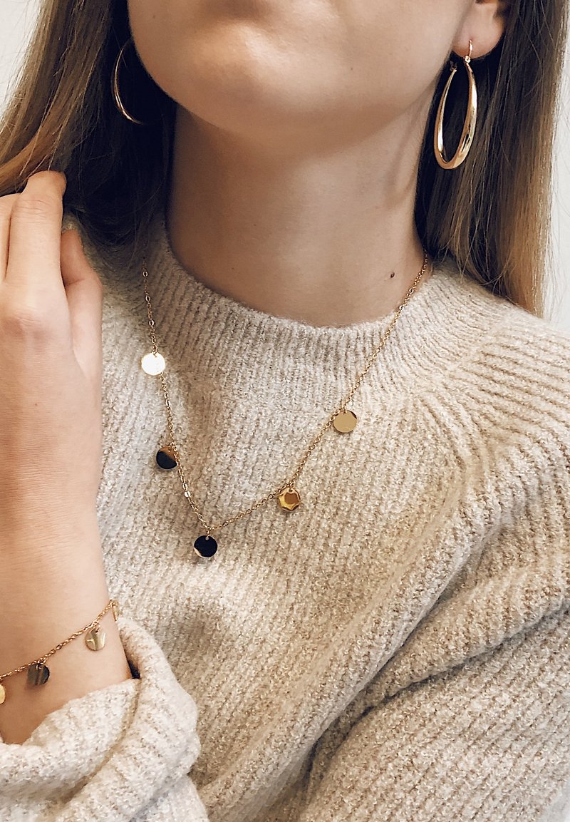 Les bijoux en ton doré comprennent un collier avec des pendentifs ronds et des boucles d'oreilles créoles assorties. Un pull en laine crème est porté.