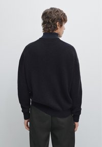 Maglione lavorato a maglia blu scuro con una texture a coste, spalle cadenti e orlo arrotondato, abbinato a pantaloni scuri e sartoriali.