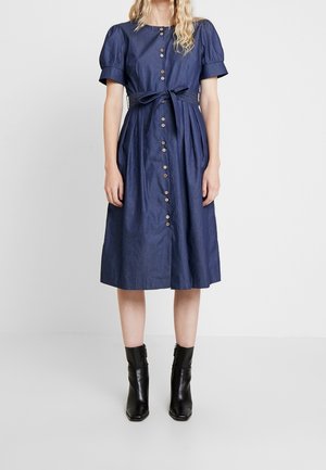 Femme portant une robe midi bleu marine avec des manches bouffantes, des boutons à l'avant, une ceinture nouée à la taille et des bottines noires.