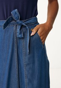 Džínová sukně s vázáním v pase, obsahující přední kapsu, světle modrou barvu a jemné švy. Hladká textura a neformální styl.