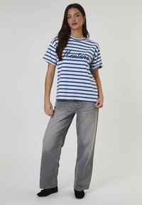 Camiseta de rayas blancas y azules con mangas cortas y un eslogan en cursiva negro. Combinada con jeans gris claro de pierna ancha y zapatos negros.