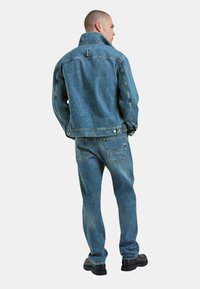 Giacca in denim blu chiaro e jeans abbinati con una vestibilità rilassata. Presenta un colletto alto, chiusura con bottoni e cuciture a vista. Calzature nere.